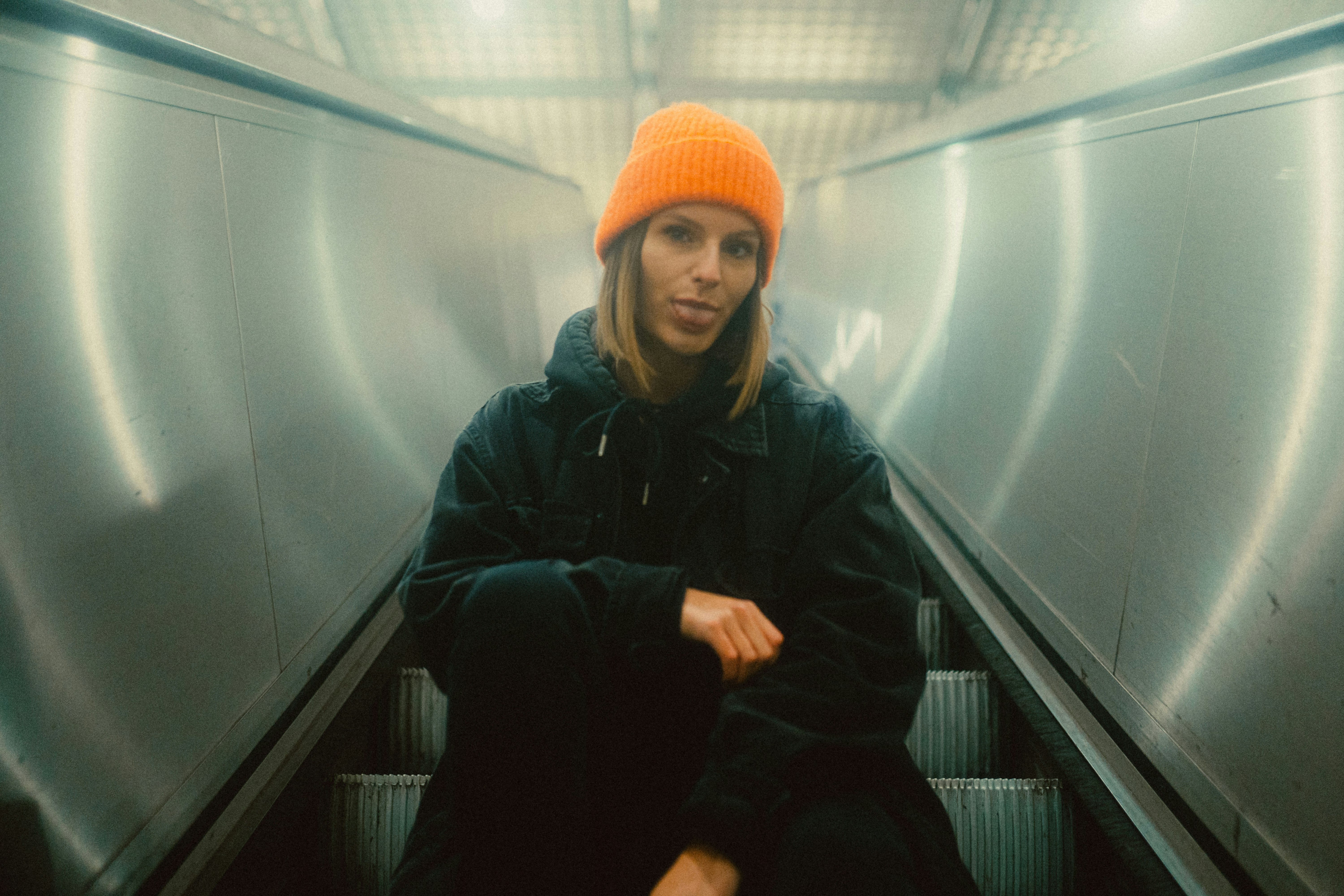woman in black jacket and orange knit cap sitting on escalator